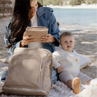 Mother holding a portable baby change mat and her child sitting on a beach with a beige nappy bag backpack beside