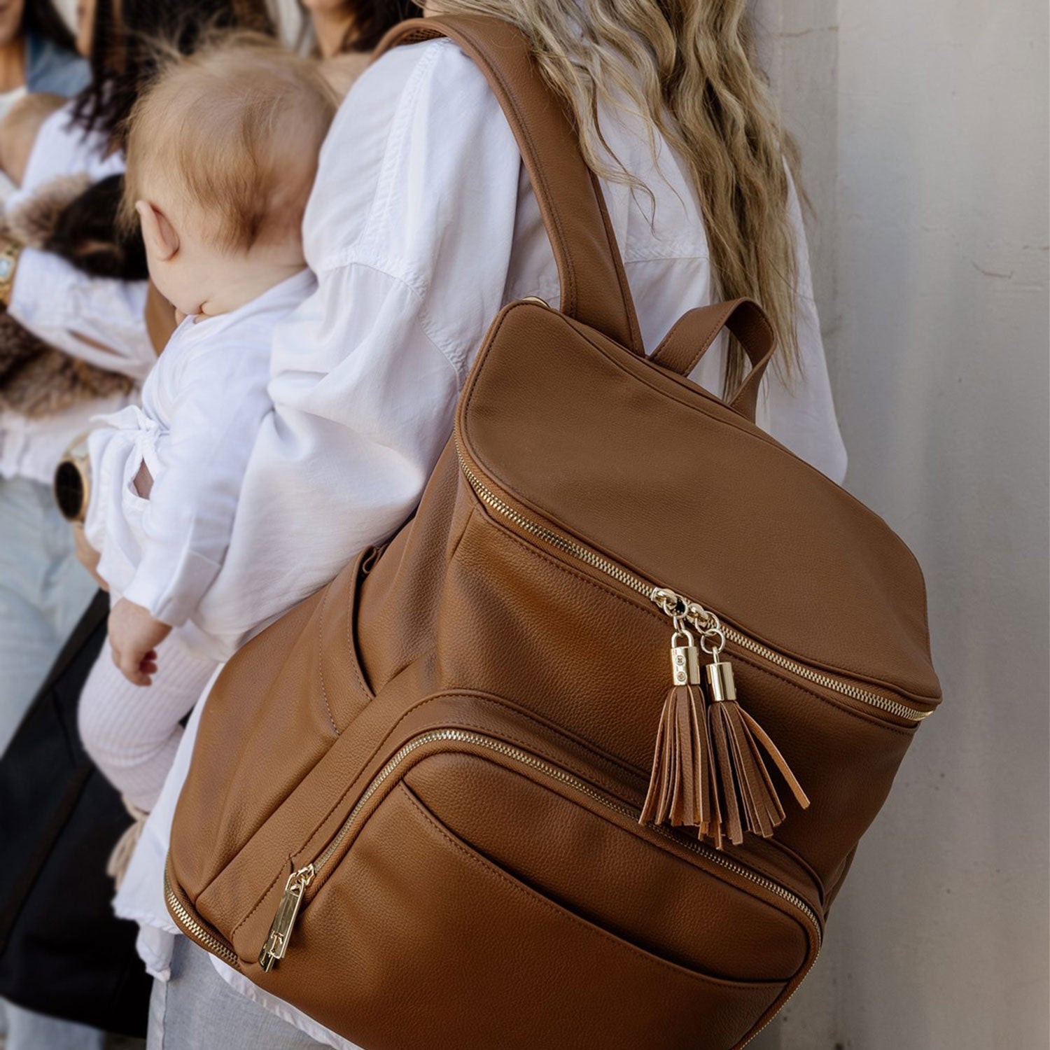 Brown nappy bag backpack with tassels worn by a pregnant woman holding a baby