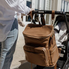 Brown diaper backpack with a stroller, held by a person wearing a white shirt and blue jeans.