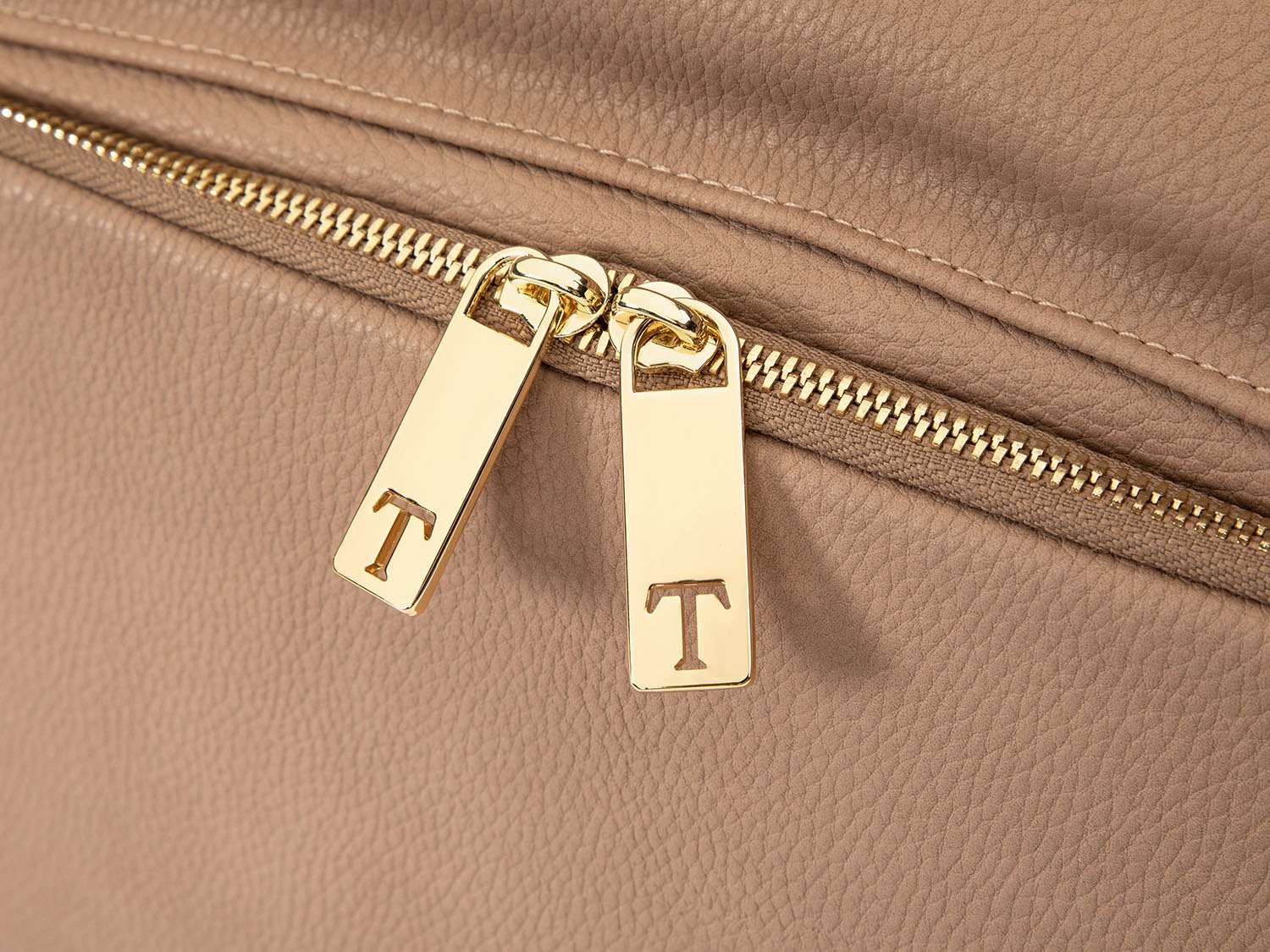 Close-up view of two gold zippers featuring the Tottie logo on a stone leather nappy backpack with a gold zipper from Tottie
