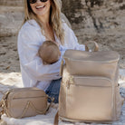 Woman holding a baby and sitting on the sea shore with a beige nappy bag backpack and a nappy crossbody bag.