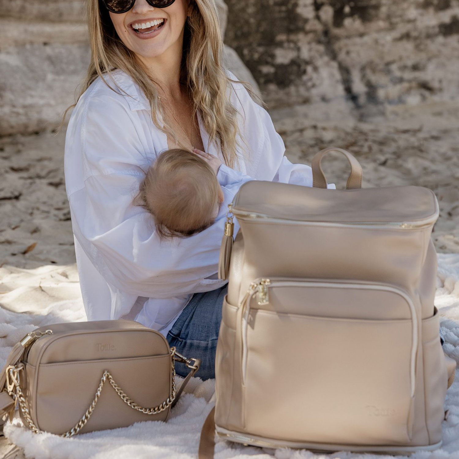 Woman holding a baby and sitting on the sea shore with a beige nappy bag backpack and a nappy crossbody bag.