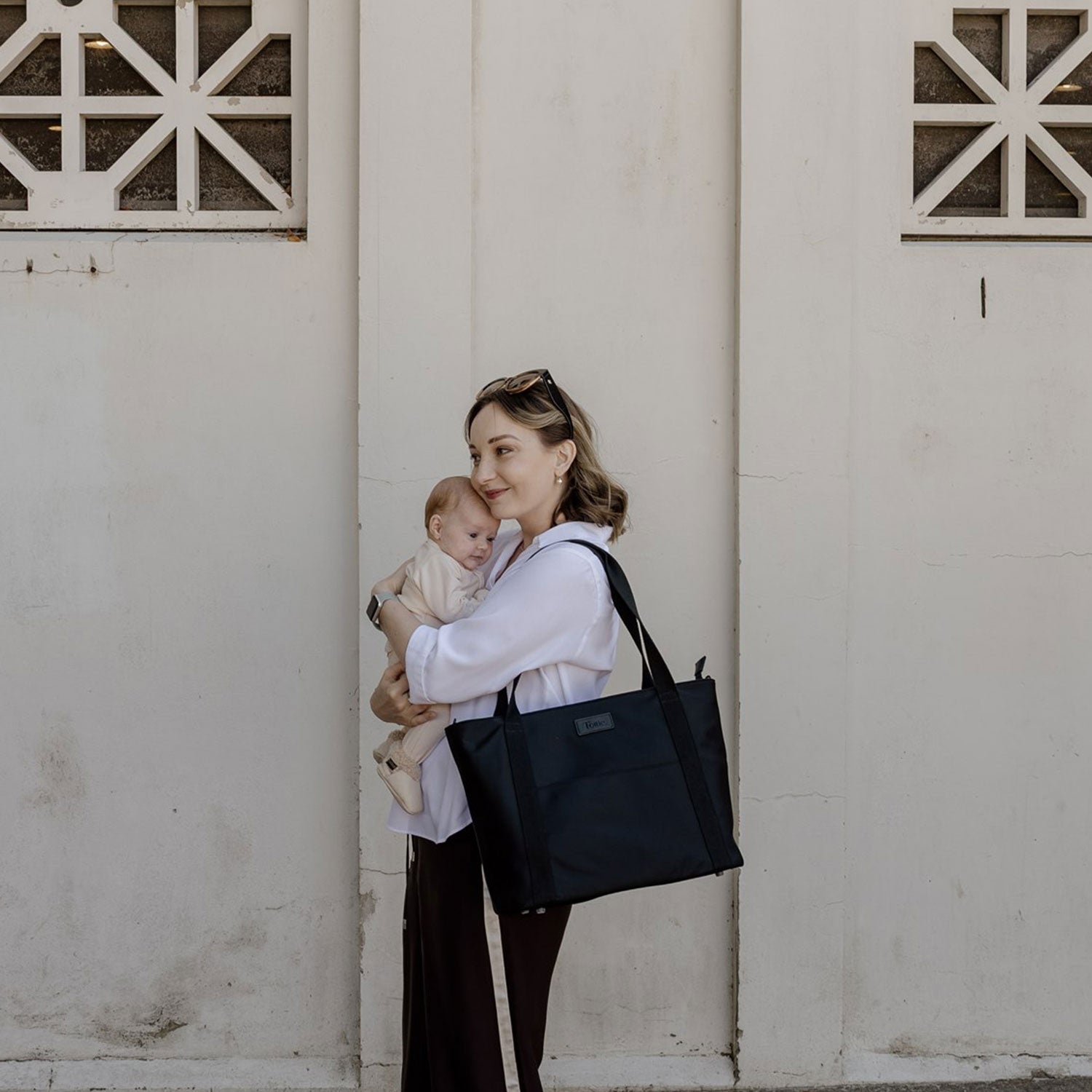 Woman holding a baby and carrying a black nylon tote bag against a white wall.