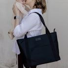 Woman holding a black nylon tote bag and a baby against a plain background