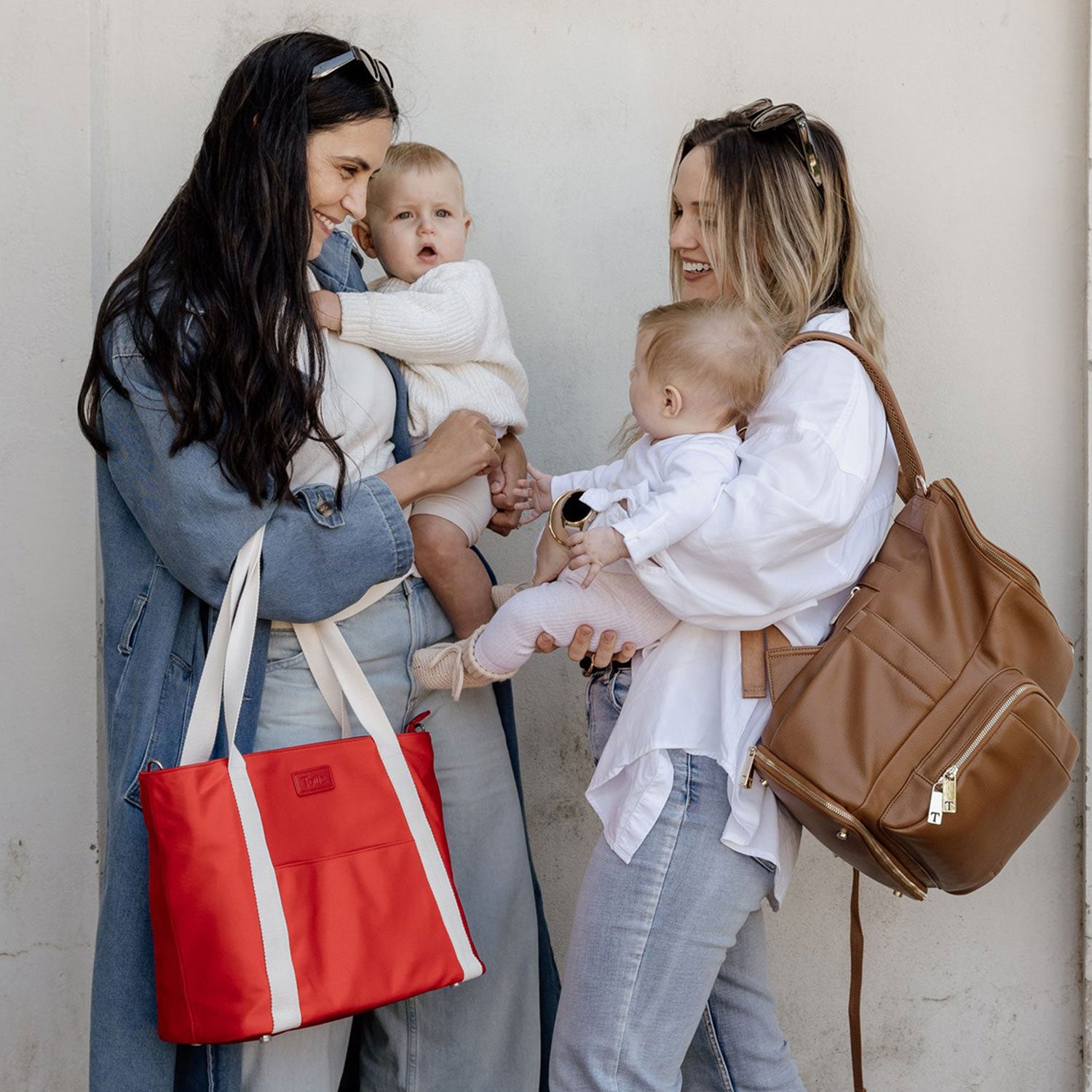 Two women holding babies and carrying nappy bags - backpack and tote against a plain background