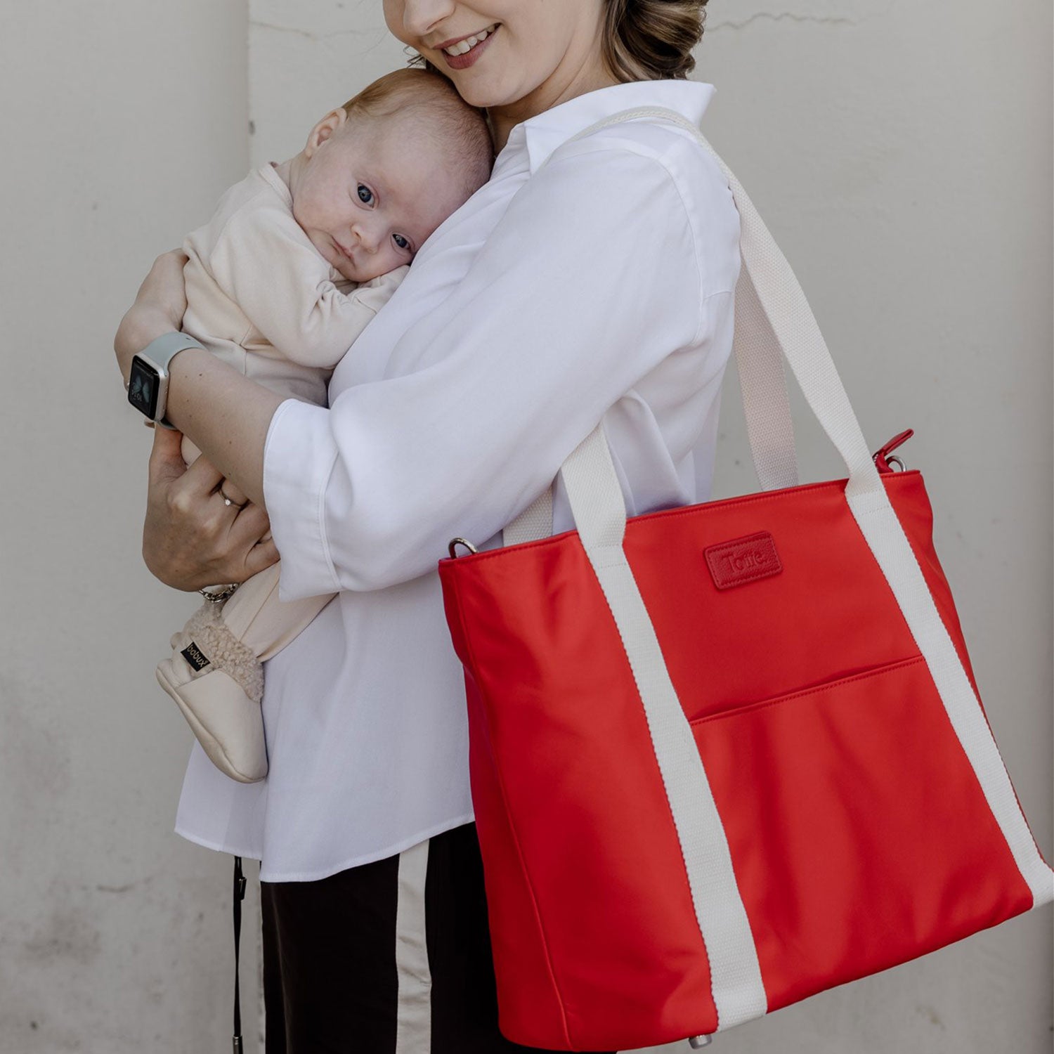 Woman holding a baby and a red tote bag with a visible brand logo.