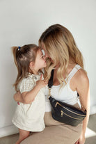 A mom carrying a black bum bag and a little girl share a sweet kiss