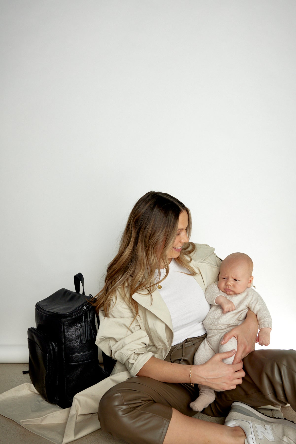 A woman sits on the floor, holding a baby, with a black travel backpack beside her