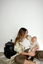 A woman sits on the floor, holding a baby, with a black travel backpack beside her
