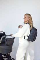 A woman wearing white pants and carrying a black travel backpack stands outdoors.