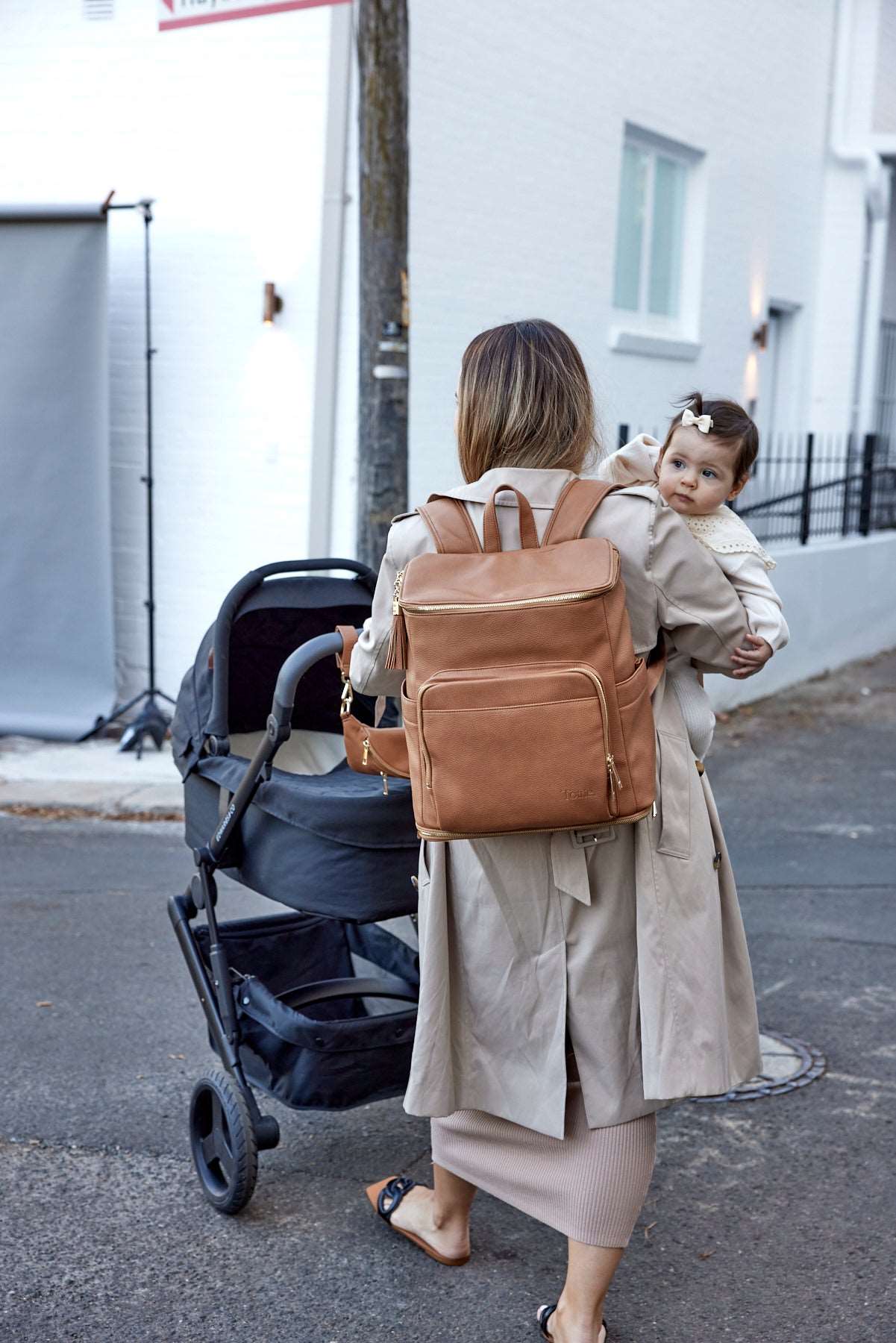 A woman pushes a stroller with a baby while wearing a tan colour backpack.