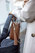 A woman pushing a stroller, with a tan colour bag with pram clips in gold hanging on the stroller