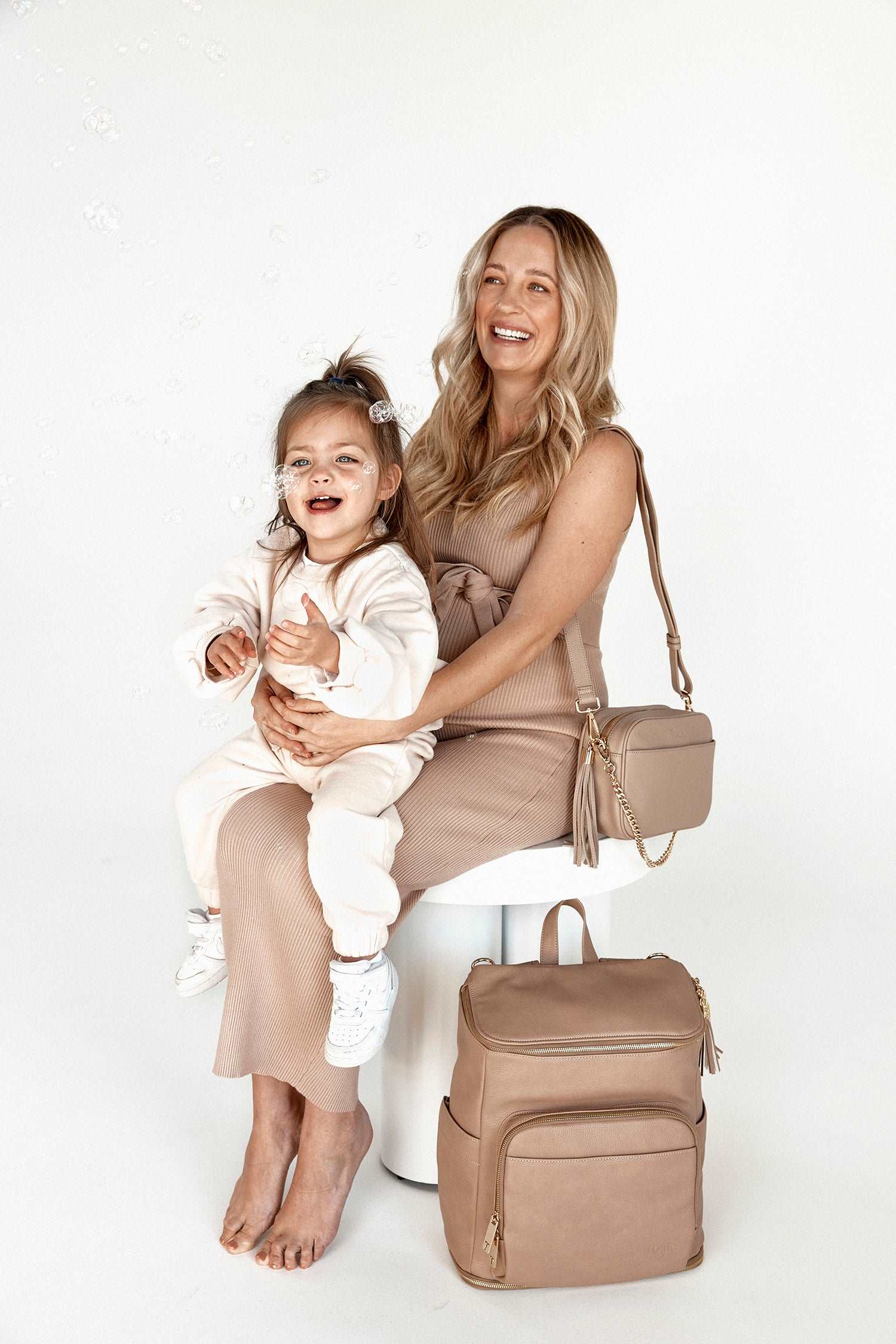 A girl sitting next to her mum, with a stone crossbody bag and a stone leather nappy backpack with a gold zipper from Tottie placed on the side.