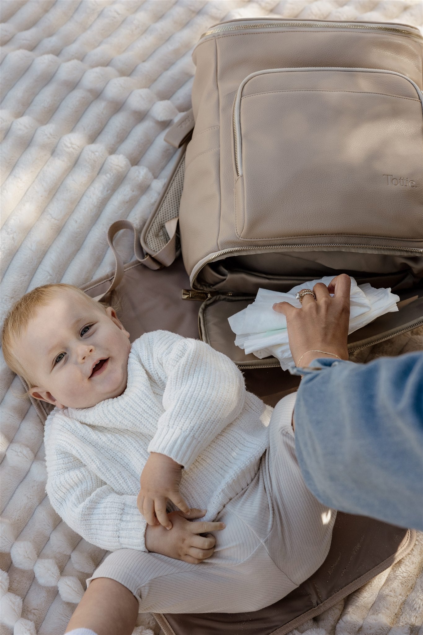Baby lying on a portable baby change mat next to an open beige nappy bag backpack with nappies, held by a mum.