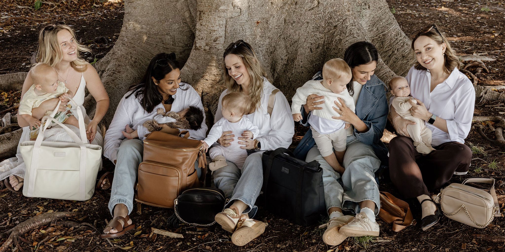Group of women and mums sitting under a tree, holding and interacting with babies, with nappy bags and travel bags displayed in front