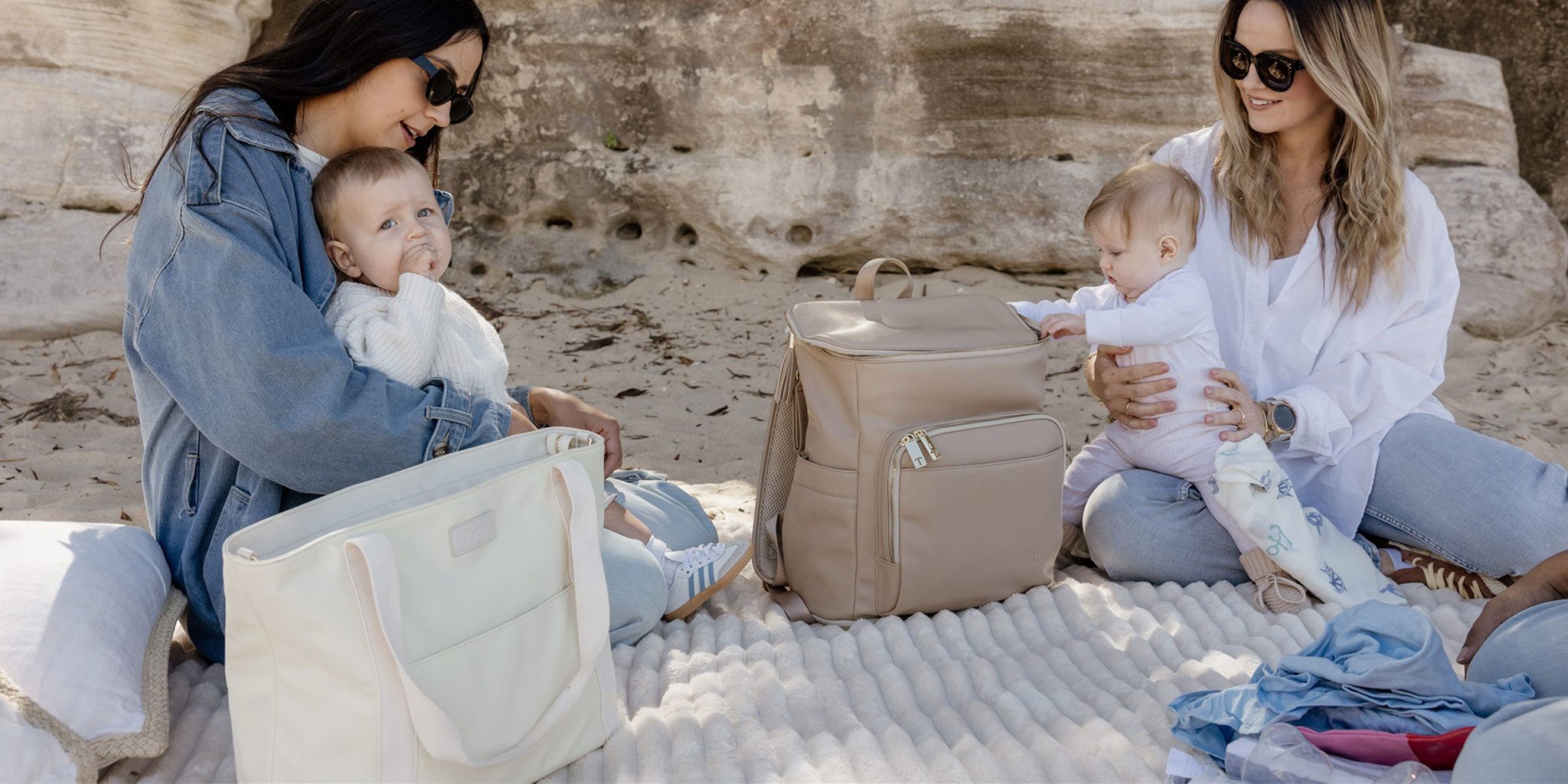Two women with babies sitting on a blanket at the beach, with nappy bags and baby items around.