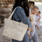 Woman wearing a pastel yellow nylon tote bag on a beach while holing her baby next to a mum holding a baby