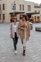 Two women stroll down a brick street, wearing women's bags from Tottie. One is wearing a stone crossbody bag, and the other is wearing a tan bum bag.