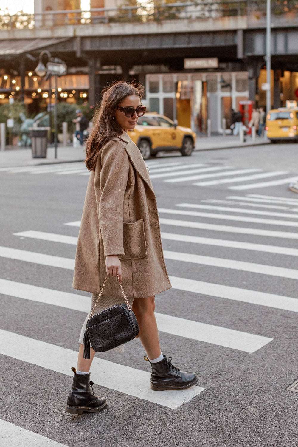 A woman wearing sunglasses, a blazer, and boots crosses the street while carrying a black Bestie Crossbody Bag from Tottie