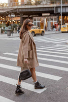 A woman wearing sunglasses, a blazer, and boots crosses the street while carrying a black Bestie Crossbody Bag from Tottie