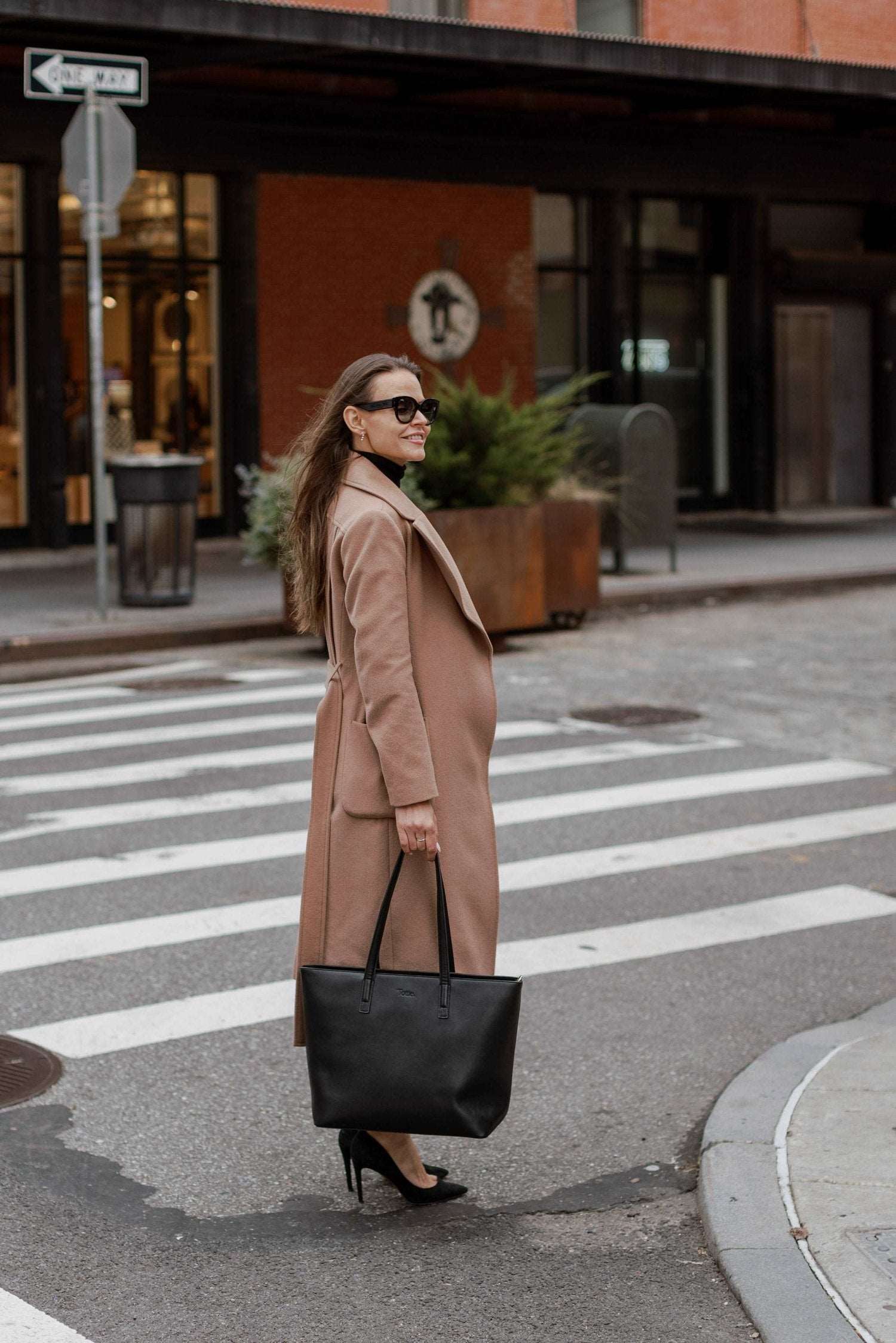 A woman in a coat and heels crosses the street while carrying a black tote bag